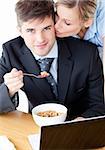 Smiling businessman eating breakfast using laptop while wife kissing his cheek