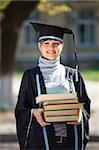 Portrait of young female graduate with stack of books, in sunlight, with blurred college building in the background