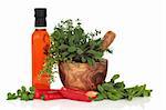 Olive chilli oil, chillies, and herb leaf sprigs of rosemary, purple and variegated sage, thyme and oregano in an olive wood mortar with pestle, isolated over white background.