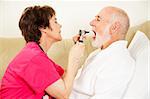 Home health care nurse uses an otoscope and tongue depressor to look in a patient's mouth.
