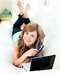 Smiling woman lying ona sofa holding a card and laptop in the living room