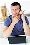 Smiling man talking on phone using his laptop in the kitchen at home