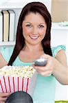 Cute caucasian woman holding a remote and popcorn looking at the camera in the living-room