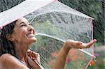 Beautiful young woman grins as she holds out her palm to catch falling water. She is holding an umbrella over her head. Horizontal shot.