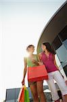 mid adult italian woman and hispanic woman carrying shopping bags out of shopping center at sunset. Vertical shape, low angle view, copy space