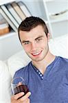 Charismatic young man holding a glass of wine sitting on a sofa in the living-room at home