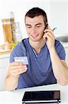 Attractive young man talking on phone holding a card in the kitchen at home