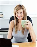 Glowing businesswoman holding a cup of coffee using her laptop at her desk in her office