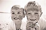 Brother and sister smile at the camera, brightly lit, on the beach, faces covered in sand.