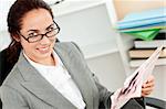 smiling businesswoman holding her glasses and reading a newspaper in her office sitting at her desk