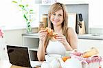 Smiling woman having breakfast in front of the laptop in the kitchen at home