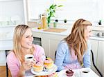 Two laughing female friends eating pastries and drinking coffee in the kitchen at home