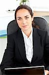 Self-assured hispanic businesswoman sitting at her desk in front of her laptop in her office