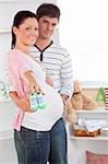 Cheerful pregnant woman holding baby shoes while husband touching her belly in the room of their future baby at home