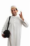 A man wearing a beautiful embroidered robe, thobe, kurta outfit fastened with ruby buttons and wearing a decorative topi hat.  He is waving in a friendly manner.  White background.