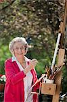 A smiling senior woman is in an outdoor setting painting. Vertical shot.