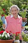 A smiling senior woman is standing in front of a table with potted flowers on a table.  She is holding a starter plant she is getting ready to pot. Vertical shot.