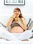 Happy beautiful pregnant woman sitting on sofa at home and eating fruit salad