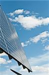 A photovoltaic solar panel array with a blue sky and puffy white clouds in the background.