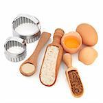 Baking products and equipment, with wholegrain flour, yeast,sugar and eggs, with wooden spoon, scoop,  and metal cookie cutters, over white background.