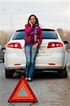 Woman calls to a service standing by a white car. Focus is on the red triangle sign. Evening light.