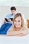 Girlfriend smiling at the camera while her boyfriend is focused on a  book sitting in the background