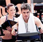 Smart female coach taking notes while her pupil is pedaling on a bicycle in a sport centre