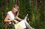 beautiful young woman sitting on  forest floor with backpack, map and bottle of water, smiling and looking in camera