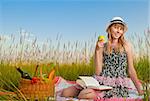 beautiful young woman having picnic on meadow, reading book, smiling and eating apple. Looking in camera, blue cloudy sky in background
