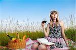 beautiful young woman having picnic on meadow, reading book, smiling and drinking wine. Looking away from camera, blue cloudy sky in background