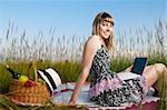 beautiful young woman having picnic on meadow, working on laptop and smiling. Looking in camera, blue cloudy sky in background