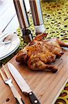 Whole barbeque chicken with cutlery and spices on wooden cutting board