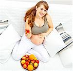 Smiling  beautiful pregnant female sitting  on sofa and  holding fruit in hand