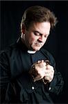 Priest holding his rosary and praying.  Black background and dramatic lighting.