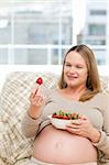 Adorable pregnant woman looking at a strawberry while relaxing on the sofa