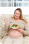 Beautiful pregnant woman holding a bowl of salad sitting in the living room