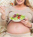 Cute pregnant woman eating a salad sitting on the sofa at home