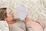 Cute pregnant woman reading a book lying on the couch at home