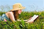 Young teenage girl reading book in summer meadow with straw hat