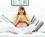 Smiling beautiful pregnant woman sitting on sofa at home with glass of juice  in hand