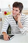 Delighted man talking on phone using his laptop in the kitchen at home