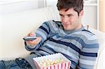 Bright young man eating popcorn and holding a remote lying on the sofa at home