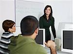 Computer class with caucasian female teacher talking to hispanic student. Horizontal shape, focus on background