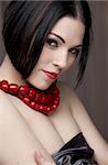 Sexy naked young caucasian adult woman with red lips, short black hair and a pierced eyebrow, covered in a dark satin sheet and wearing a red coral necklace