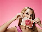 mid adult blond woman holding photo of her mouth kissing on pink background. Horizontal shape, front view, head and shoulders, copy space