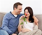 Lovely man looking at his girlfriend smelling a red rose together on the sofa