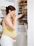 italian 6 months pregnant woman standing near refrigerator and eating fat food. Waist up, vertical shape, side view