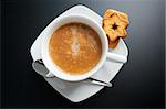 White porcelain cup of freshly brewed coffee top view close-up arranged with two sandwich-biscuits, spoon and plate on dark background