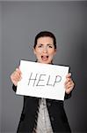 Stressed business woman imploring for help, holding a cardboard with the message "Help". Focus is on the cardboard