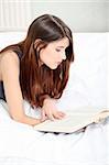 Beautiful young brunette woman reading a book on the bed, at home